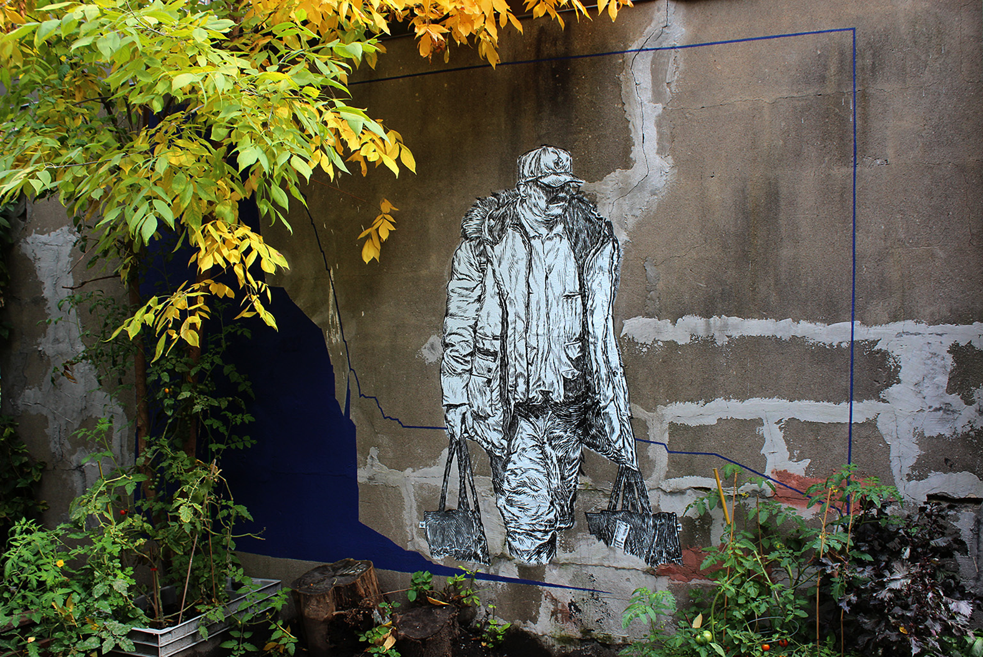 A black and white street art mural of a man walking with bags, set against a textured gray wall, partially surrounded by greenery.