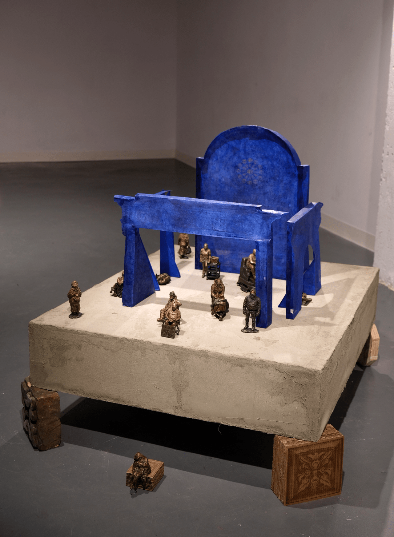 A mixed-media art installation featuring a large blue arch structure on a square concrete base, surrounded by small bronze figurines depicting seated figures.