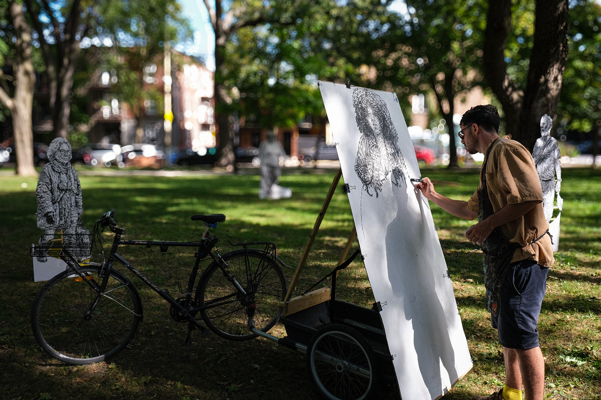 An artist drawing a portrait on a large canvas in a park, with a bicycle and surrounded by large paper cutouts of people.